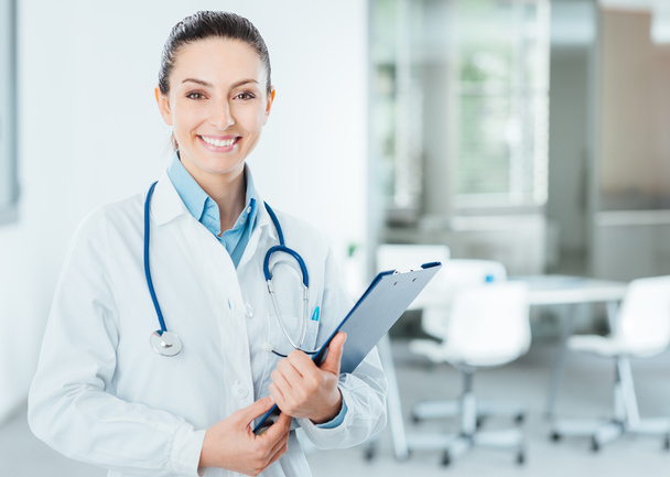 Smiling female doctor with lab coat in her office holding a clipboard with medical records, she is looking at camera